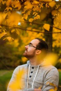 An autistic person stands in a park, looking up contemplatively through the trees